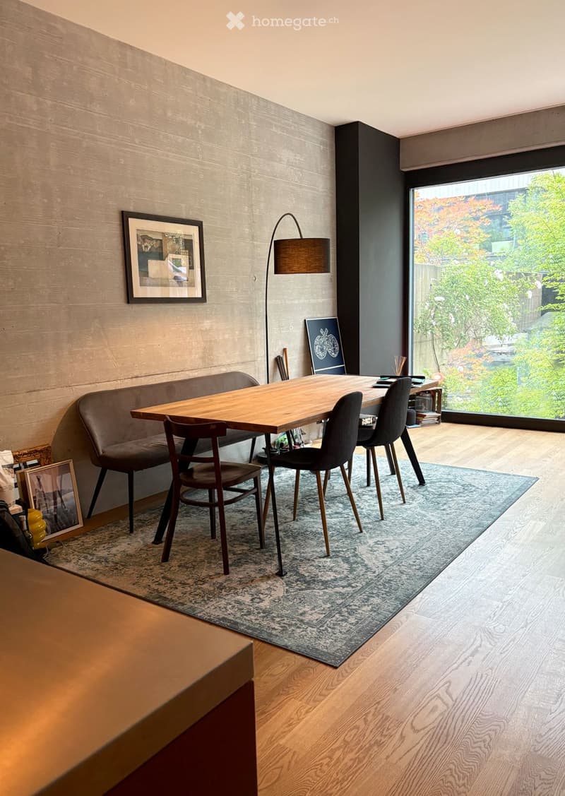 Modern dining area with a wooden table, four chairs, a grey sofa, a floor lamp, and a large window overlooking a garden.