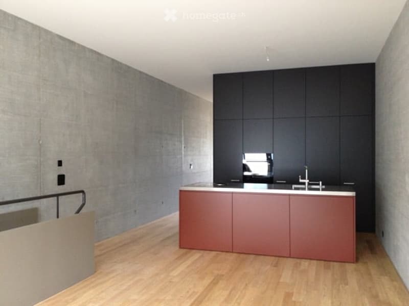 Modern kitchen with a red island, black cabinets, and a sleek faucet. The space features light wooden flooring and a concrete accent wall.
