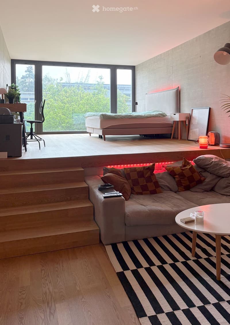 Modern, multi-functional room with a raised sleeping platform, sofa, coffee table, and large windows offering a view of greenery.