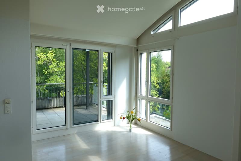Bright, empty room with large windows and sliding glass doors leading to a balcony. Light-colored wooden flooring and white walls.