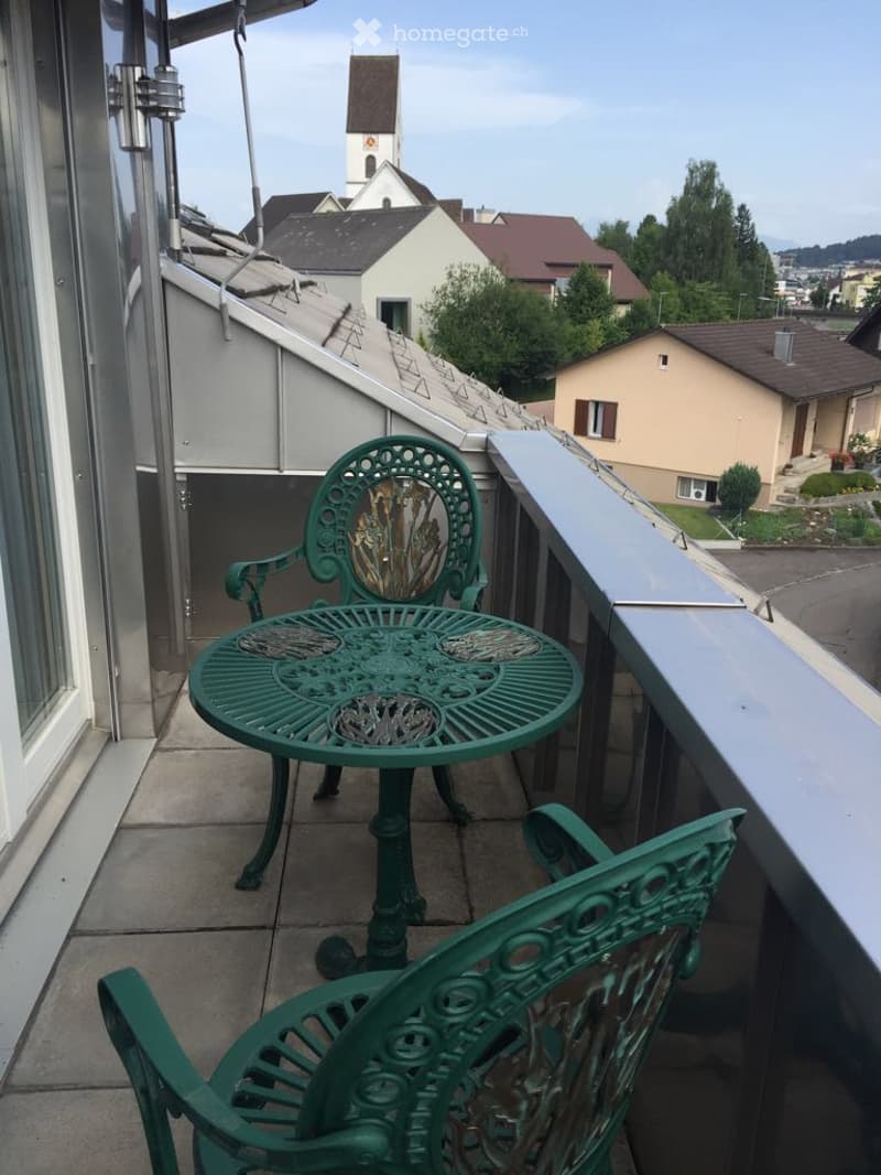 Small balcony with green metal table and chairs, concrete floor, railings, view of surrounding buildings and trees