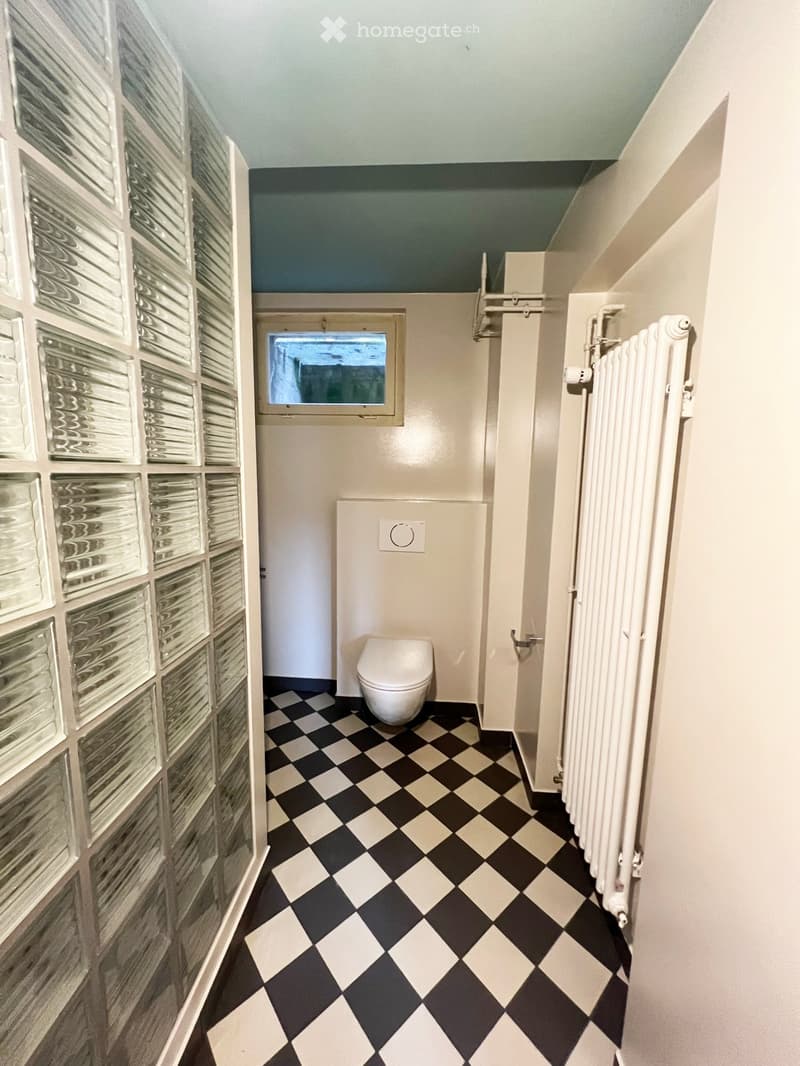 Bathroom with checkered black and white floor, white toilet, frosted glass block wall, radiator, and small window.