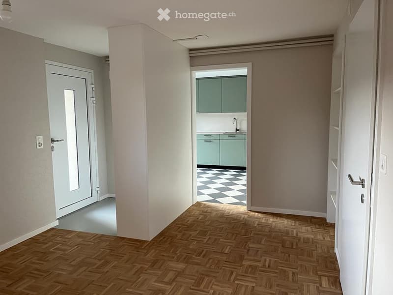 Empty room with wooden flooring, a door, and a small kitchen area visible in the background. The kitchen features mint green cabinets and a checkered floor.