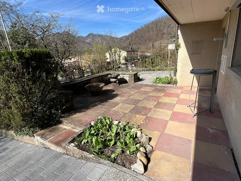 Balcony with tiled flooring, small garden bed with plants and rocks, stone benches, and a scenic mountain view.