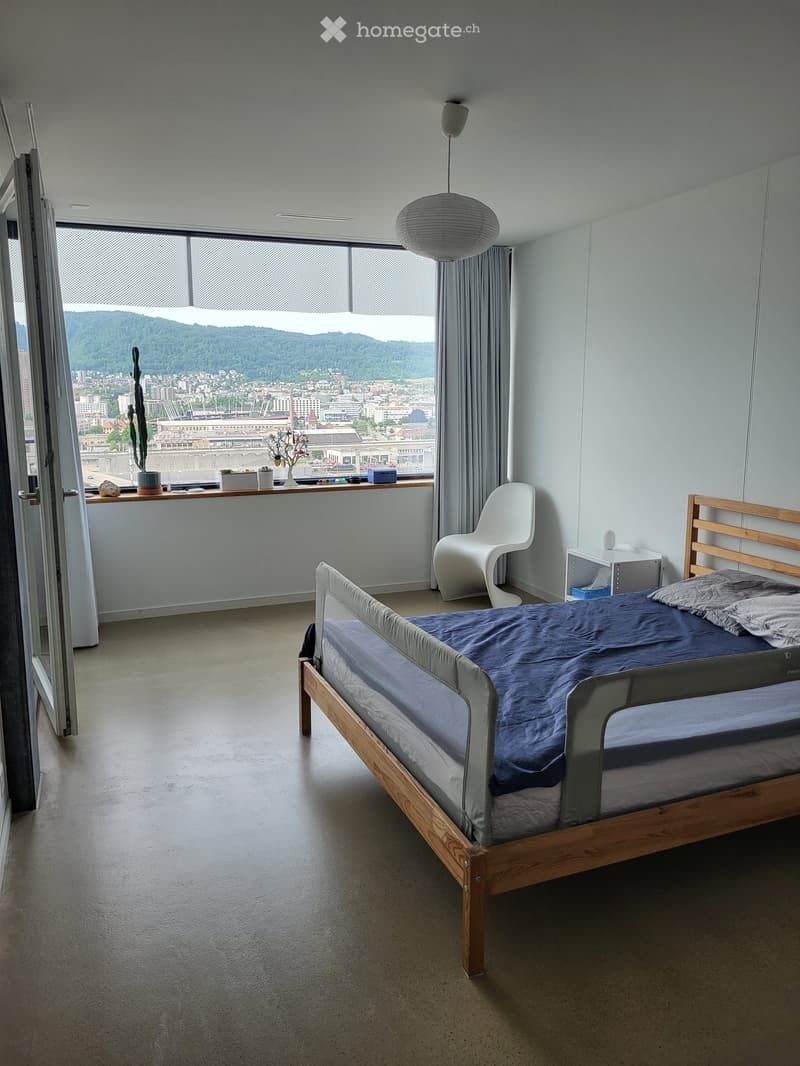 Modern bedroom with a wooden bed, blue bedding, white chair, nightstand, and a view of the city and mountains through large windows.