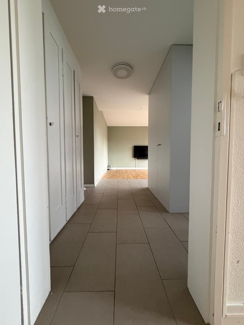 Empty hallway with tiled floor, white walls, multiple doors, and a visible room with a TV at the end.