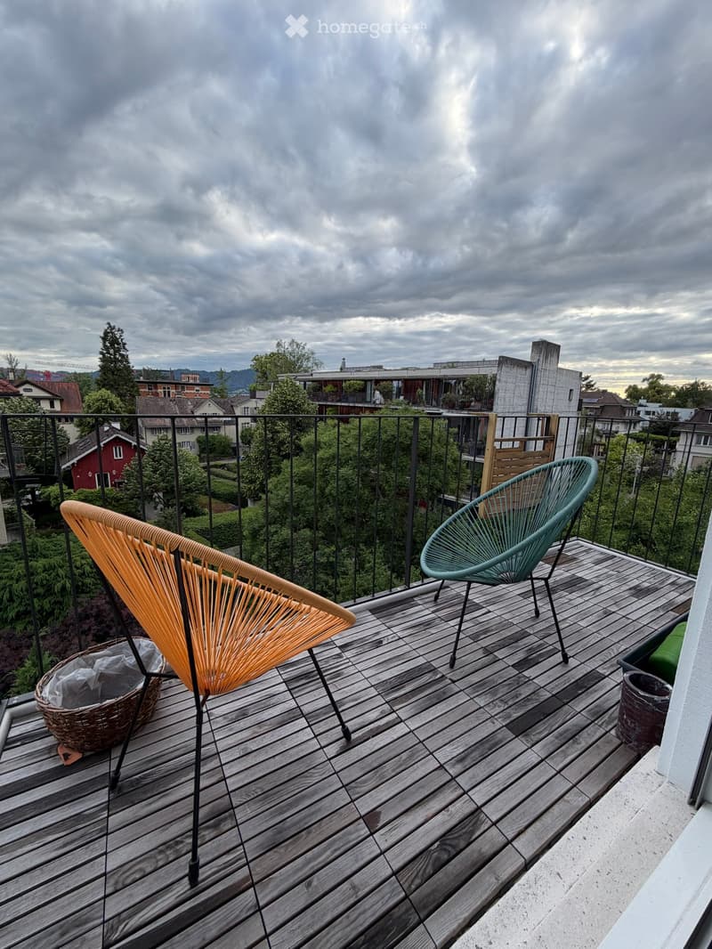 Wooden deck with two modern chairs, a wicker basket, and a view of the city and greenery.