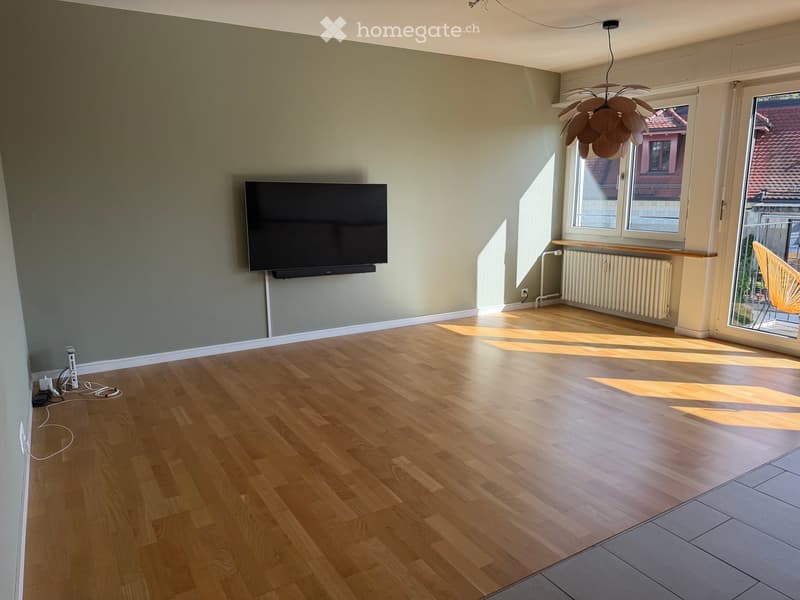 Empty room with wooden flooring, large windows, and a mounted TV on the wall. Natural light streams in, and there is a radiator and a hanging light fixture visible.