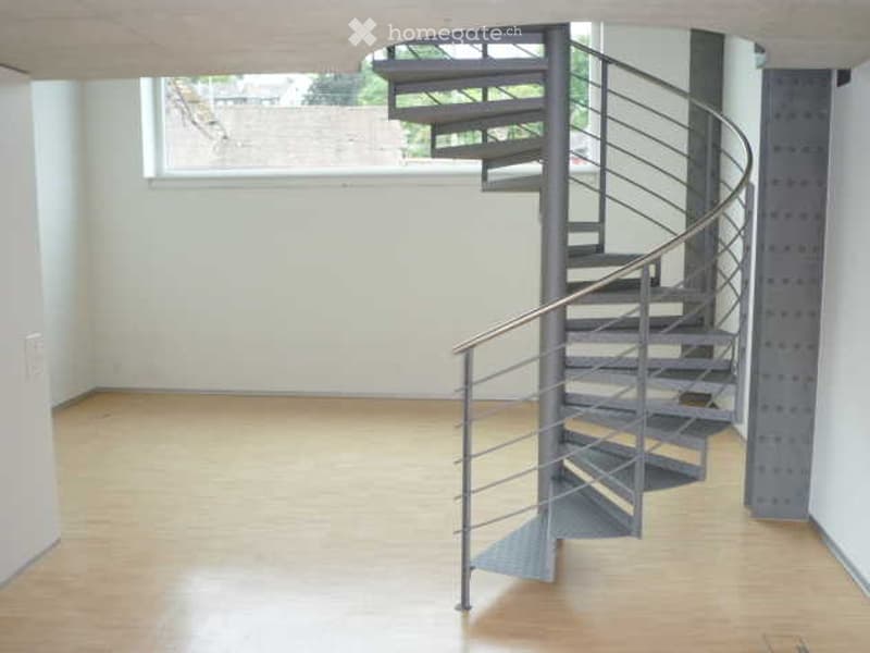 Empty room with wooden flooring and a modern spiral staircase leading to an upper level. The walls are white, and there is a large window on one side.