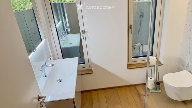 Modern bathroom with a white bathtub, sleek faucet, toilet, and a large mirror. The room features wooden flooring and a glass-enclosed shower area.