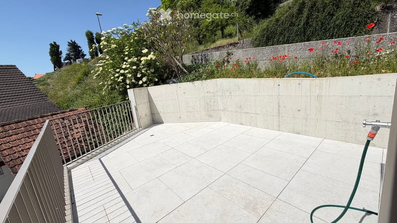 Clean, tiled balcony with metal railing, concrete walls, and a garden visible beyond.