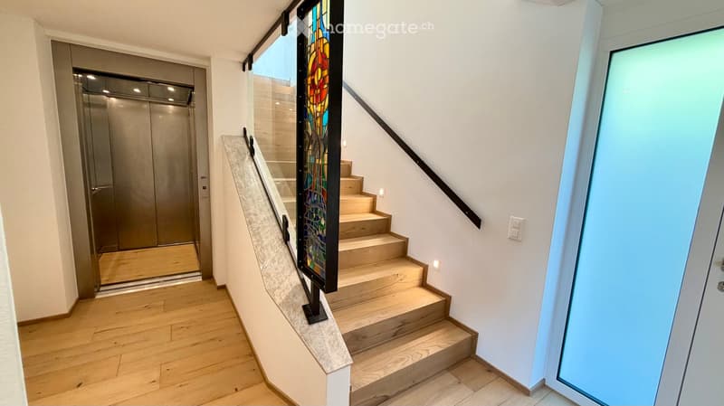 Modern hallway with wooden flooring, a staircase with a colorful stained-glass railing, and an elevator on the left side.