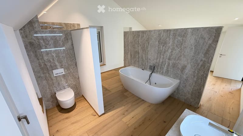 Modern bathroom with wooden flooring, a large bathtub, toilet, and sink. Features marble walls and recessed lighting.