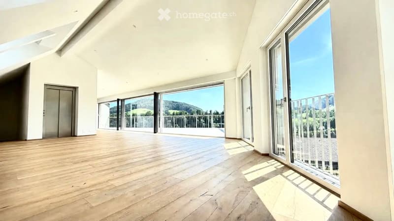 Bright, empty room with wooden flooring, large glass sliding doors leading to a balcony with scenic views, white walls, and a high ceiling.