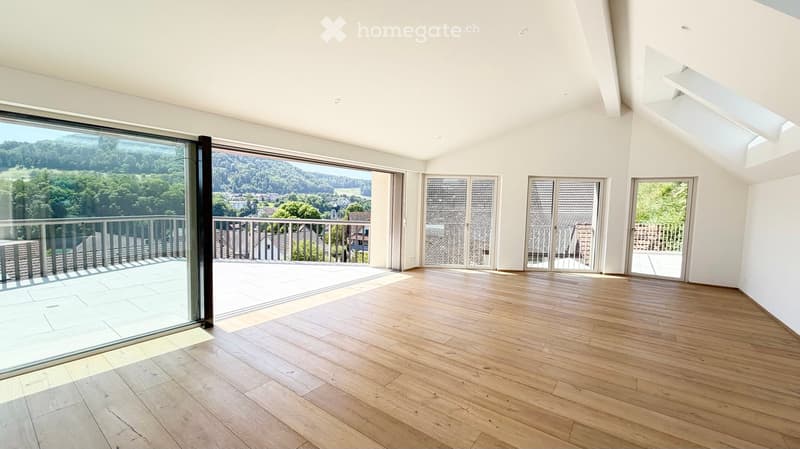 Bright, empty room with wooden flooring, large glass doors leading to a balcony with a scenic view of trees and a town.