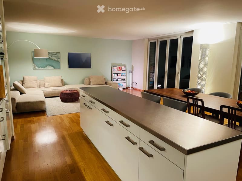 Modern open-plan living and dining area with a sectional sofa, a round wooden stool, a kitchen island, a dining table with chairs, and a floor lamp. The space features wooden flooring, large windows, and contemporary decor.