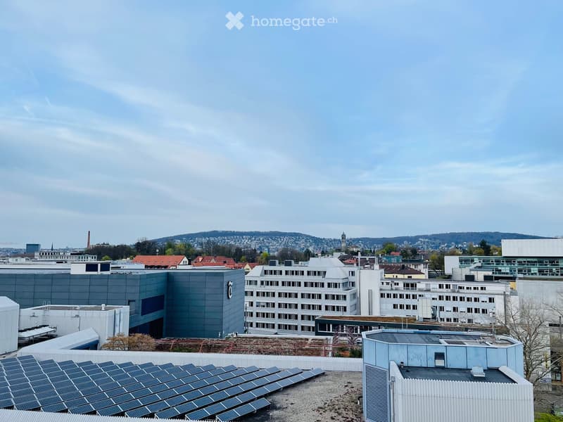 A panoramic view of a cityscape with multiple buildings, including modern structures and older architecture, under a partly cloudy sky. Solar panels are visible on a rooftop in the foreground.