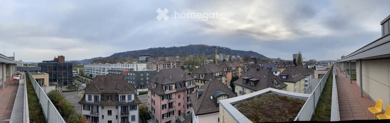 Balcony overlooking a cityscape with residential buildings, a green roof, and distant hills.