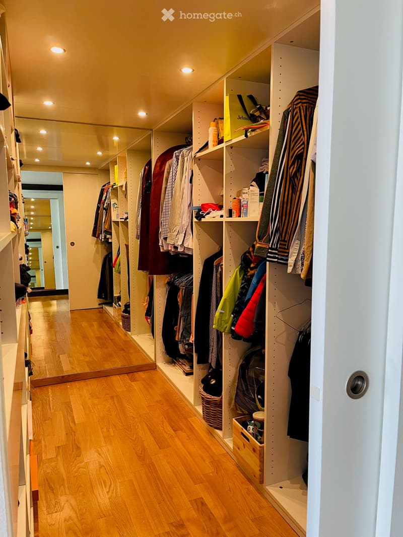 Spacious, well-lit hallway with wooden flooring, built-in closet units filled with clothing, shoes, and accessories. Features recessed ceiling lighting and a large mirror on one wall.