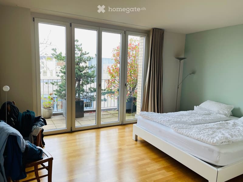 Bright bedroom with wooden flooring, large sliding glass doors leading to a balcony, a white bed with pillows and duvet, a wooden chair with clothes draped over it, and a floor lamp near the balcony.