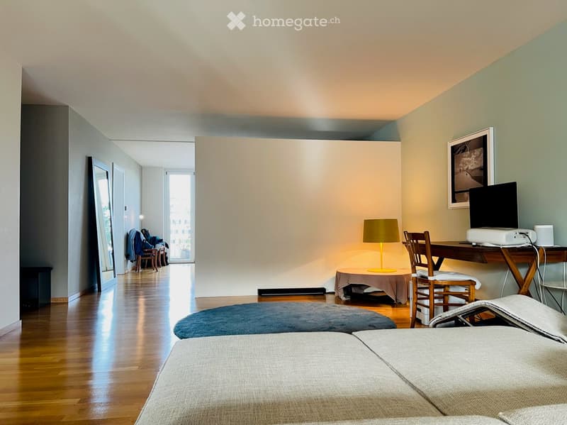 Modern bedroom with two single beds, a desk with a chair, a lamp, and a screen. The room features wooden flooring and large windows.