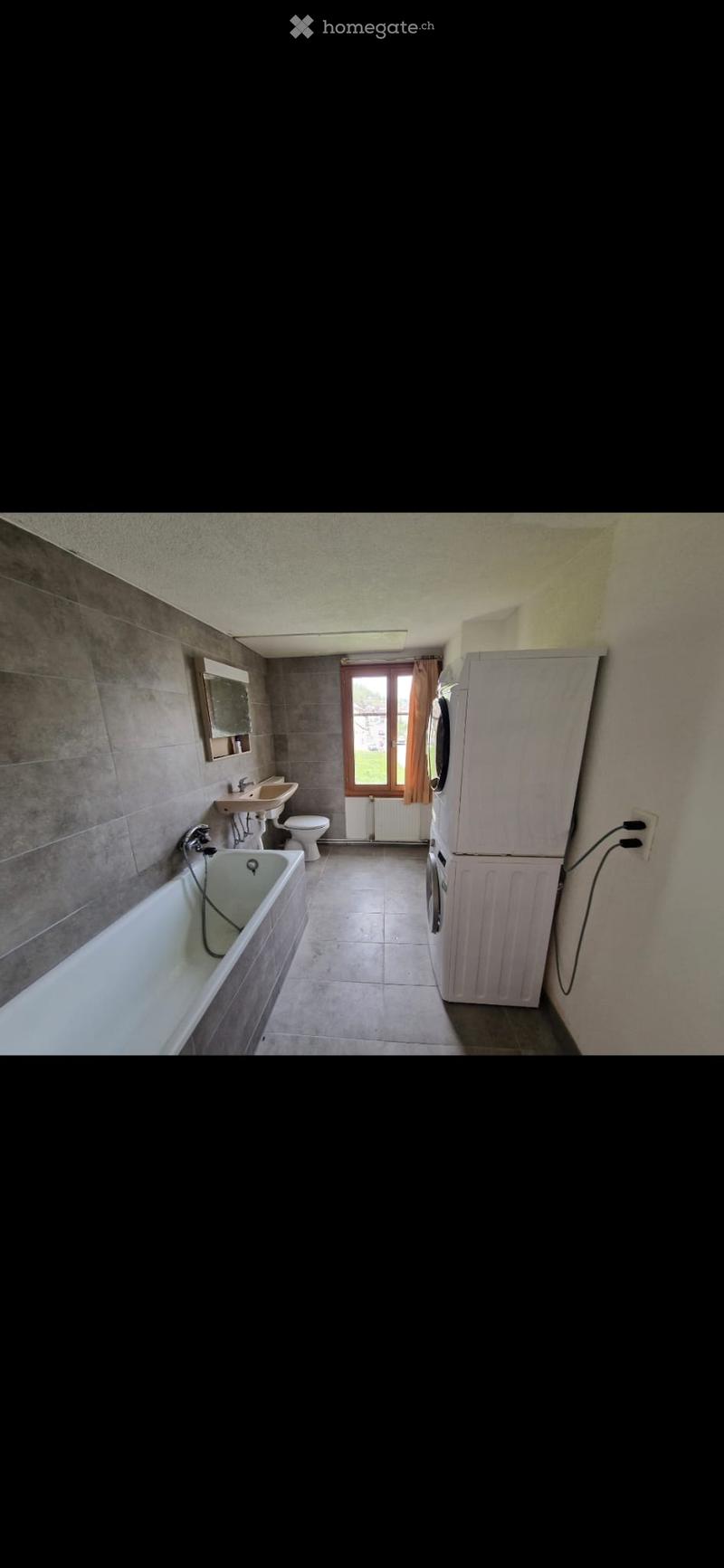 Bathroom with a bathtub, sink, toilet, mirror, and a washing machine. The room has tiled walls and a window with curtains.