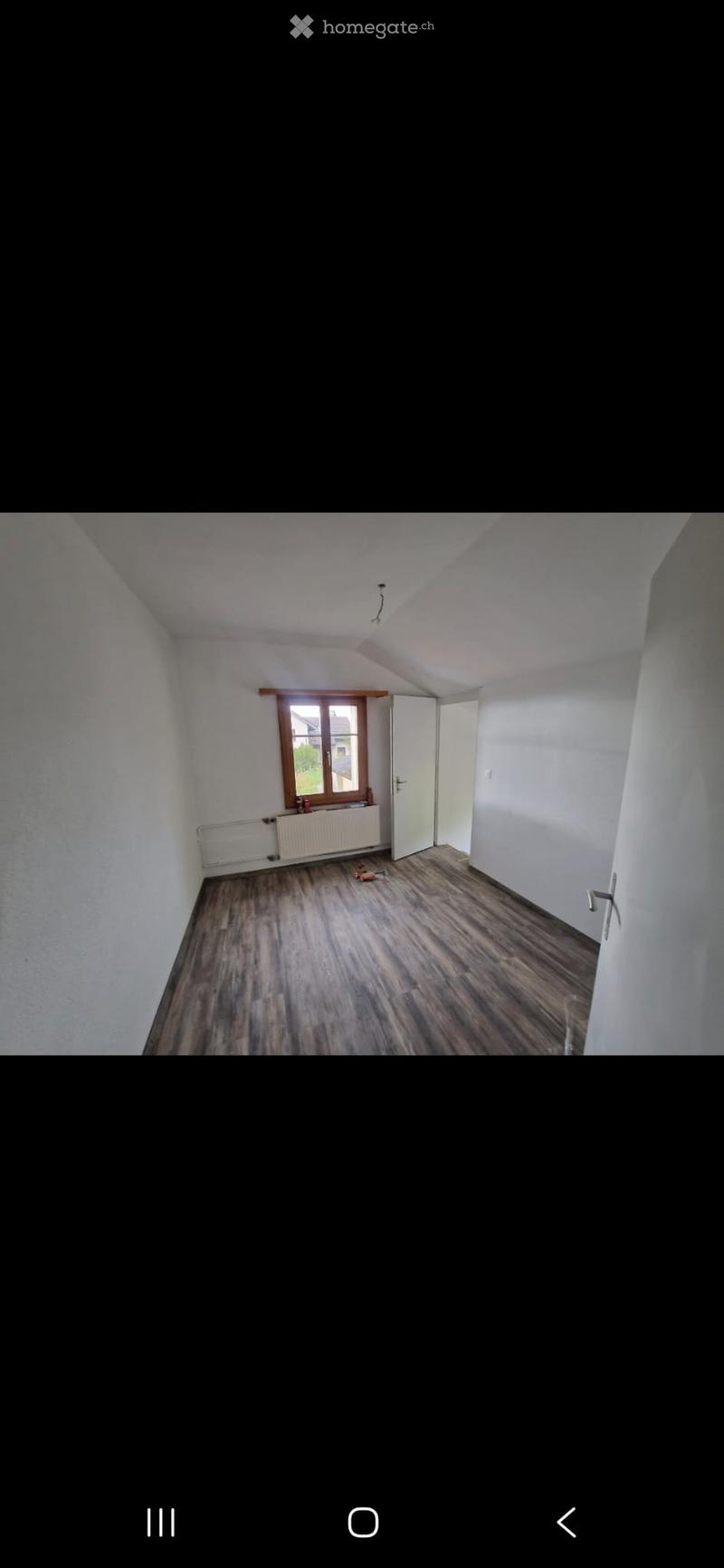 Empty room with wooden flooring, white walls, a window with wooden frame, a radiator, and a door leading to another space.