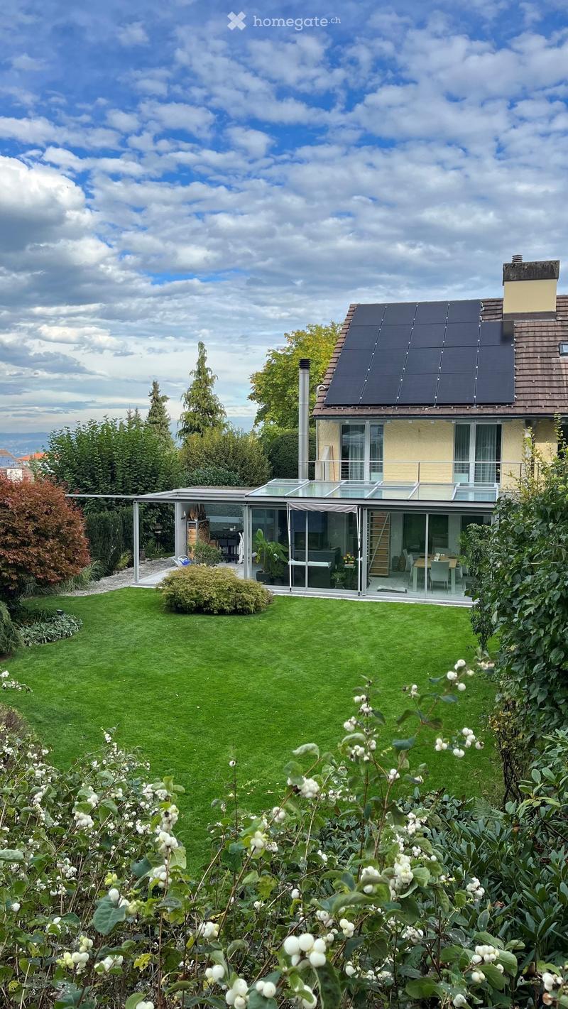 A well-maintained garden with a lawn, various plants, and a modern glass extension attached to a house. The extension includes a dining area with chairs and a table. The house has solar panels on the roof and is surrounded by trees and shrubs.