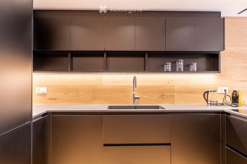 Modern kitchen with dark brown cabinets, wooden backsplash, stainless steel faucet, and countertop appliances including an electric kettle and coffee maker.