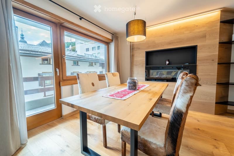 Modern dining area with a wooden table, four chairs, a table runner, a television, and a hanging lamp. Large windows offer a scenic view of the town.