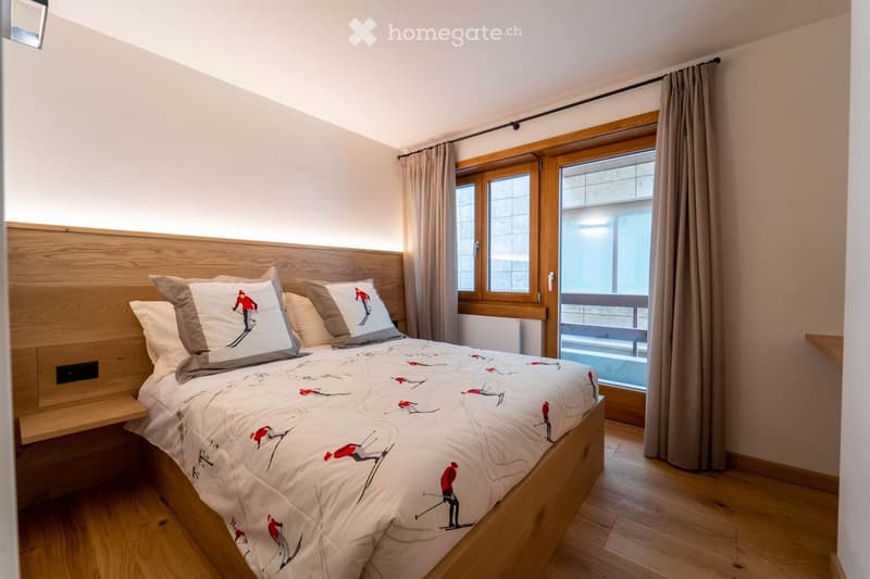 Cozy bedroom with wooden flooring, large bed with a ski-themed duvet and pillows, wooden headboard, and large windows with curtains.