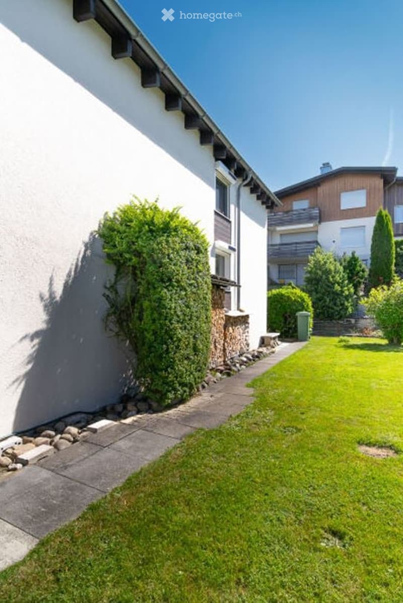 Well-maintained garden area with a neatly trimmed lawn, a concrete pathway, and a large evergreen bush near the house wall. The house has a white exterior with dark roof trim and multiple windows.
