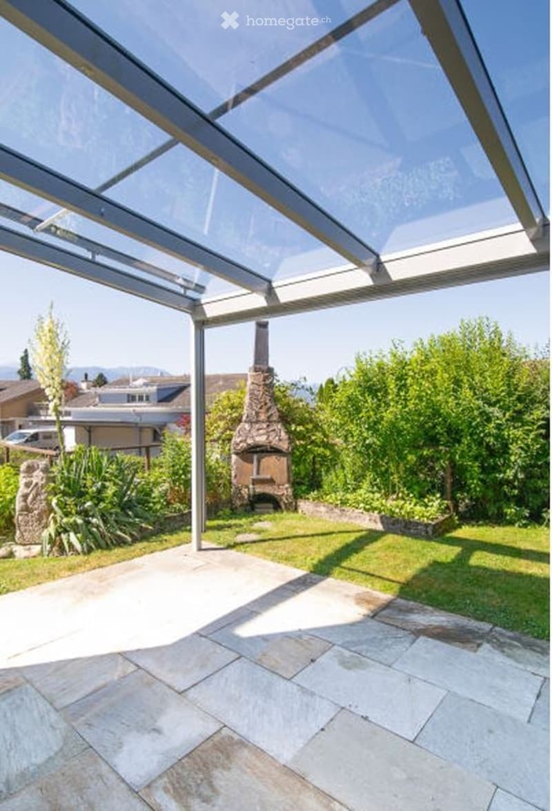 A spacious outdoor patio with a stone fireplace, surrounded by lush greenery and a clear glass roof structure.