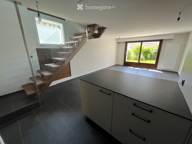 Modern, unfurnished kitchen with sleek grey cabinets, dark tiled floor, and a large window overlooking a green area. The space includes a central island and a staircase leading to an upper level.