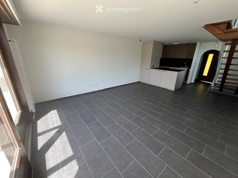 Empty room with dark grey tiled floor, white walls, and a small kitchen area with white cabinets and a sink.