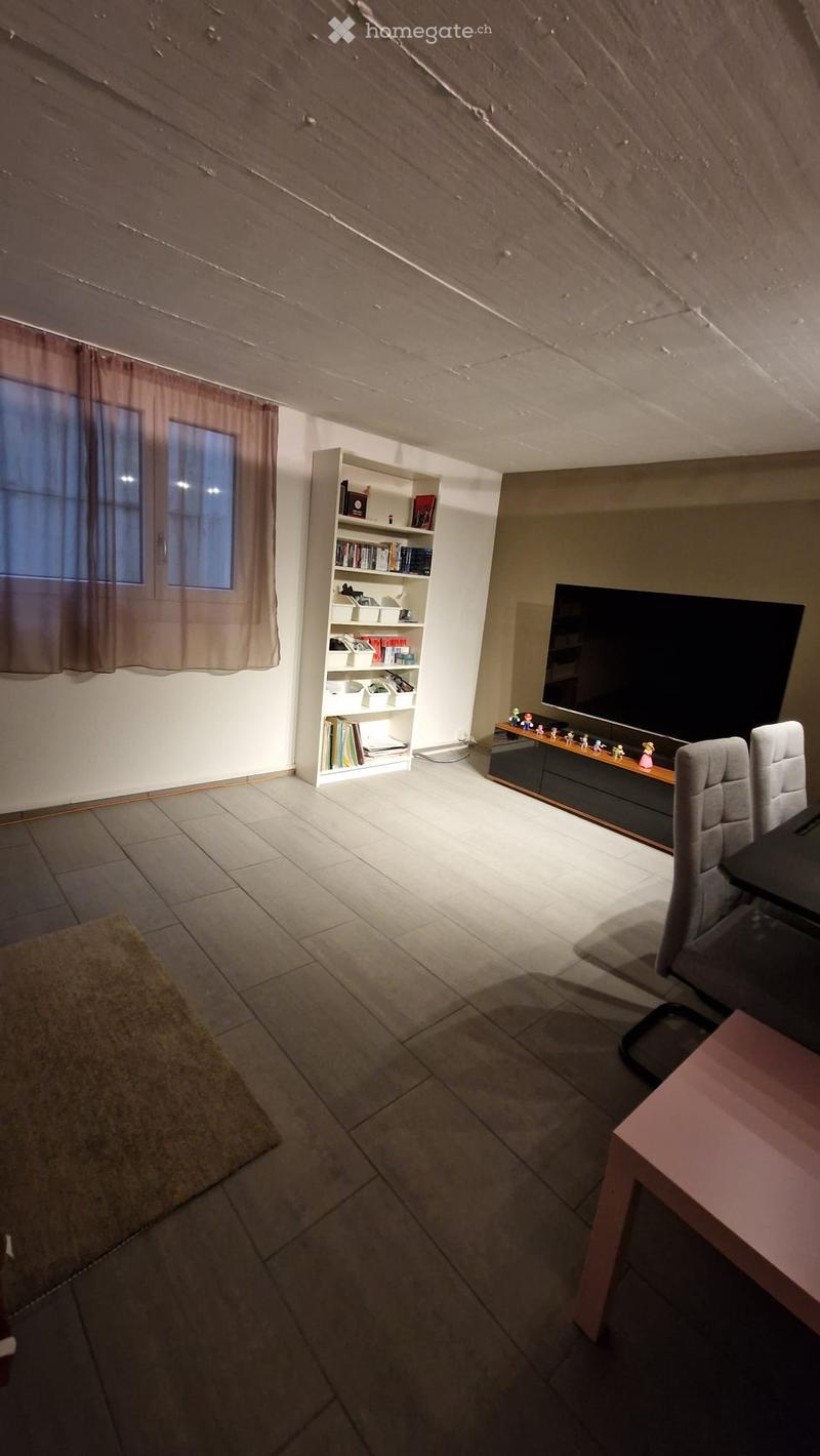 A furnished room with a tiled floor, a large window with curtains, a shelf with books and decorative items, a television on a stand, and a small table with chairs.