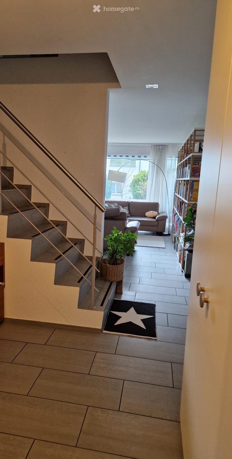 Modern hallway with tiled floors, staircase with metal railing, and a view into a living area with a sofa, bookshelf, and plants.