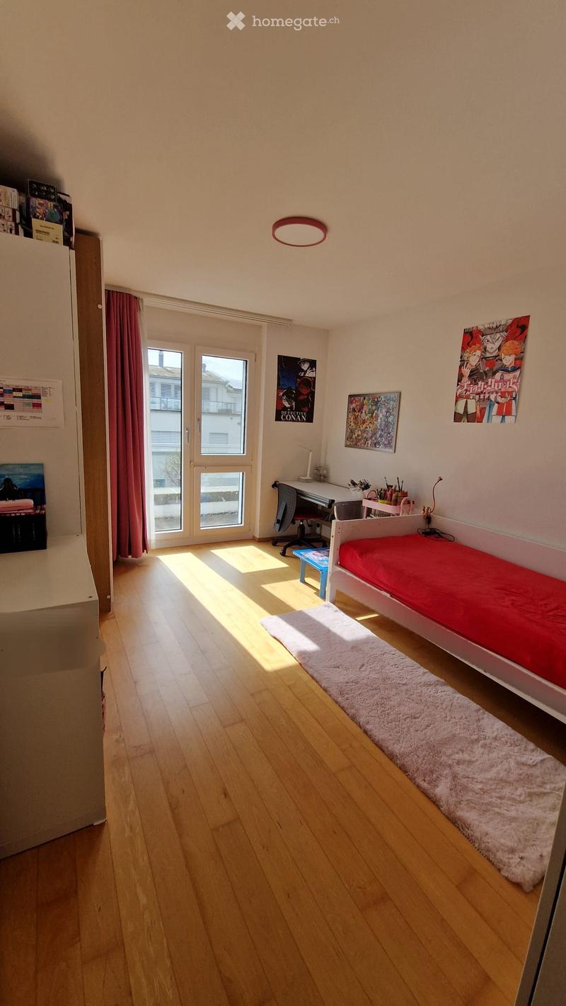 Bright bedroom with wooden flooring, a single bed with red bedding, a desk with a chair, and a window with red curtains. Artwork and posters adorn the walls.