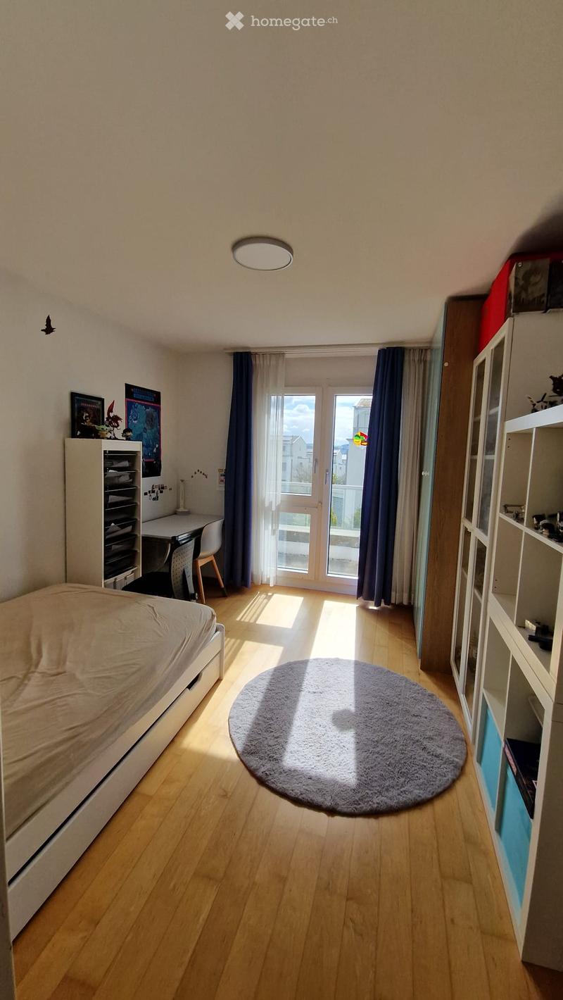 Bright bedroom with wooden flooring, a single bed, a round rug, a desk with a chair, and shelves with various items. Large window with curtains leads to a balcony.