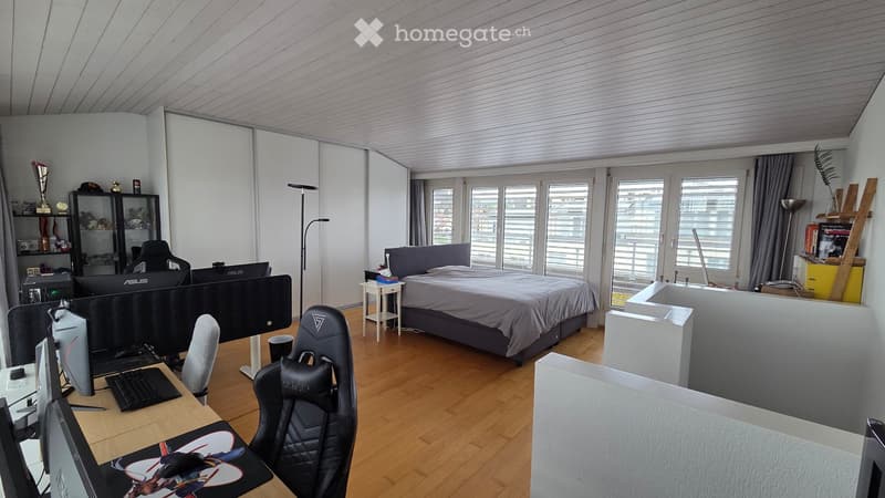 Furnished Room. A multi-functional room with a bed, desk setup, and gaming chair. The space features a wooden floor, large windows, and modern furniture.