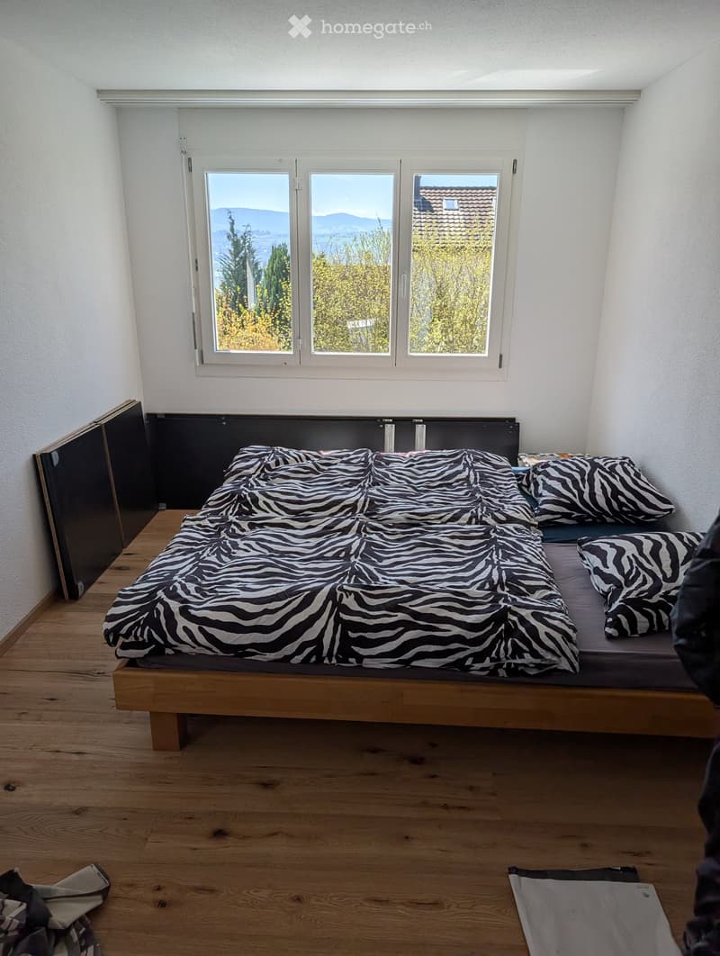 A modern bedroom with a wooden floor, large window offering a scenic view, and a neatly made bed with a zebra-patterned blanket and matching pillows.