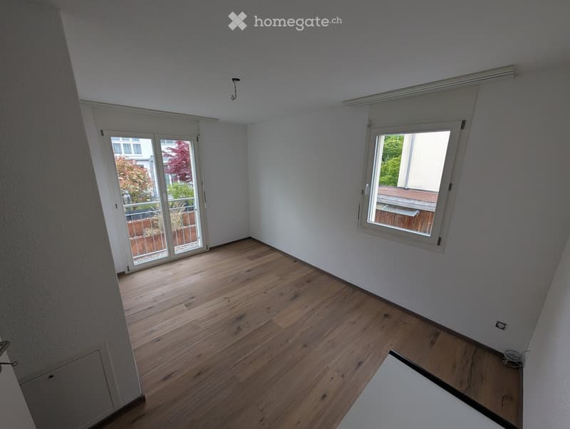 Empty room with wooden flooring, white walls, and large windows offering views of the outdoors.