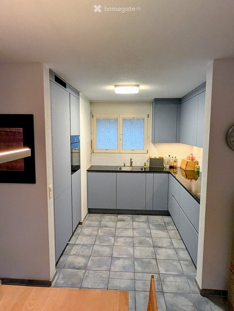 Modern kitchen with grey cabinets, tiled floor, sink, and various bottles on the counter.