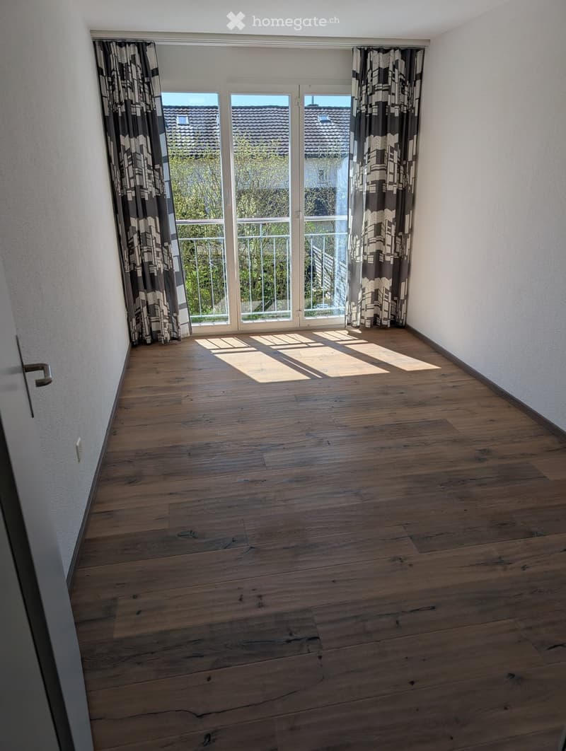 Empty room with wooden flooring, large window with curtains, and a door leading to a balcony.