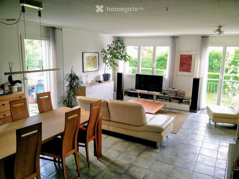 Bright living room with large windows, cream-colored sofa, wooden coffee table, dining table with chairs, TV, speakers, and decorative plants.
