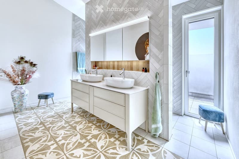 modern bathroom, double sinks, white cabinets, herringbone tile wall, tile floor, stool, vase with flowers, towel hanging