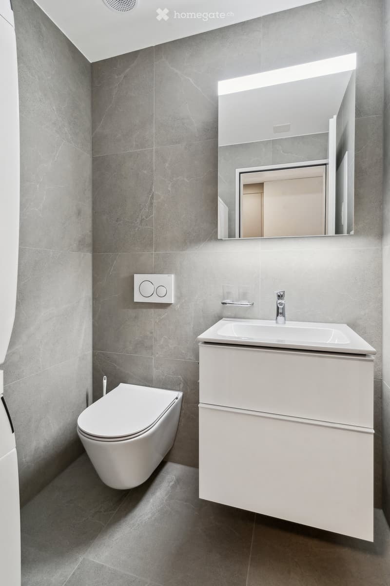 Modern bathroom with grey tiled walls, white toilet, white sink with cabinet, and a large mirror with built-in lighting.