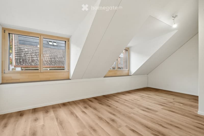 Bright, empty attic room with wooden flooring, white walls, and large windows offering views of neighboring rooftops.