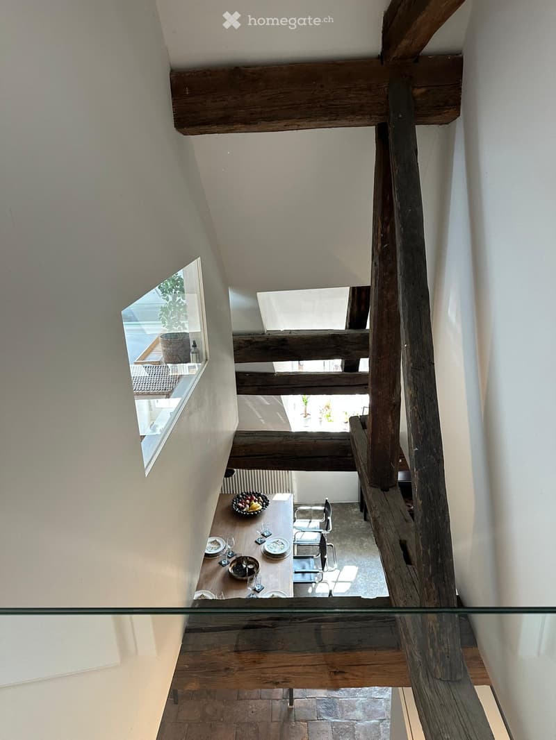 A modern, open-concept hallway with exposed wooden beams and a glass railing, leading to a dining area with a table and chairs.