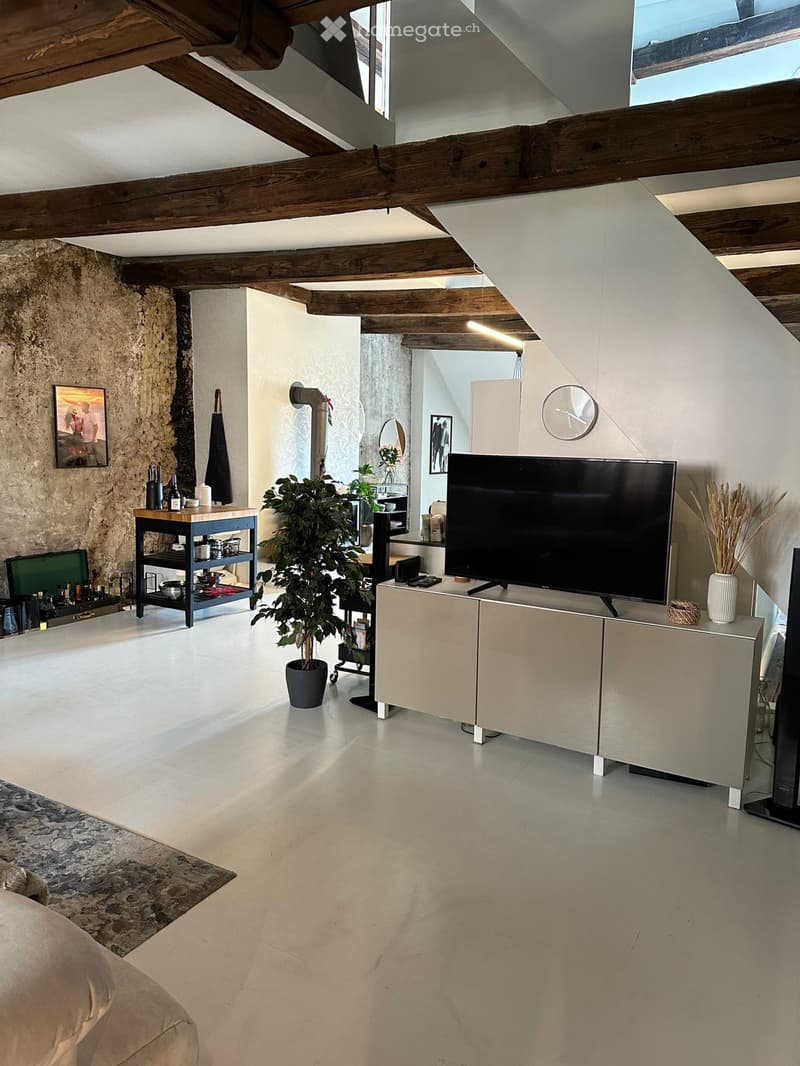 Modern living room with exposed wooden beams, stone walls, a large television on a cabinet, a small table with bottles and a plant, and a clock on the wall.